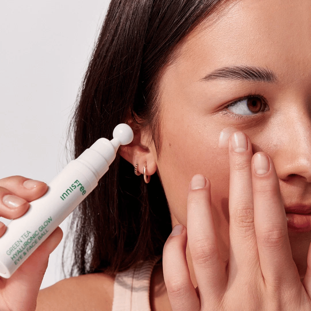Osoba aplikuje Innisfree Green Tea Hyaluronic Glow Eye & Face Ball na svoju pokožku pod očami. Produkt má bielu, okrúhlu aplikačnú časť a je v bielej fľaši so zeleným textom. Tvár používateľa je čiastočne viditeľná a zameriava sa na aplikáciu produktu.