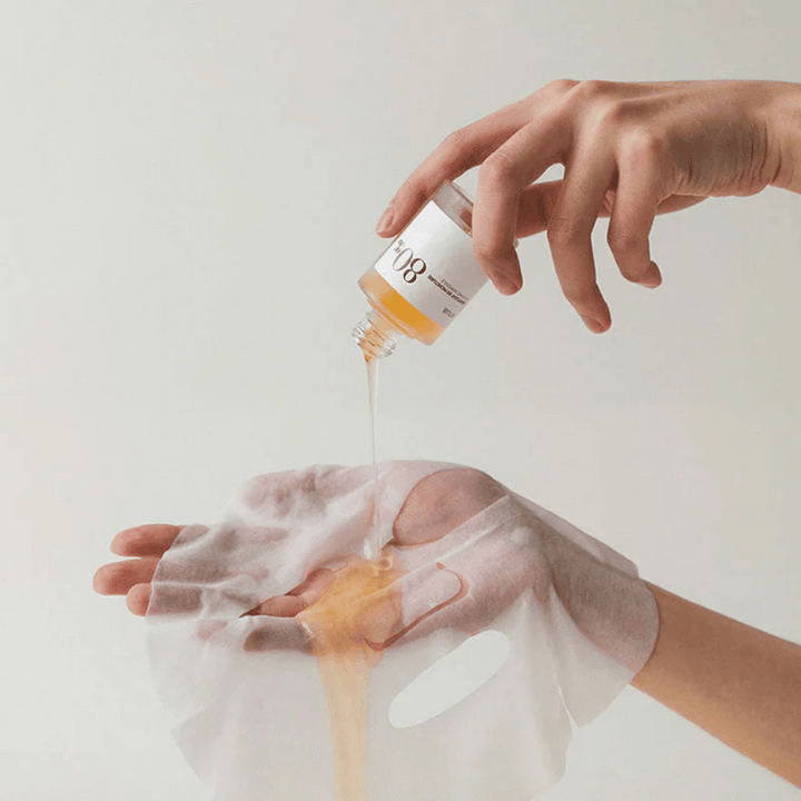 Ruka drží zlatú, hustú tekutinu z malej fľaštičky označenej '108' na priehľadnú pleťovú masku, ktorú drží v druhej ruke. Pozadie je neutrálne a jednotné, aby zdôraznilo produkt.