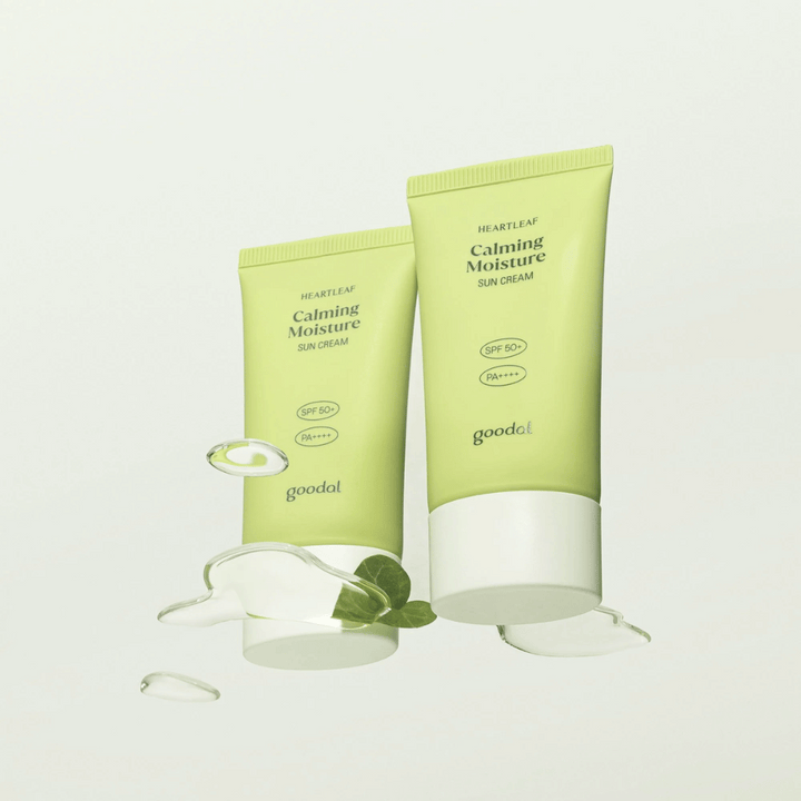 Dve tuby Goodal Heartleaf Calming Moisture Sun Cream SPF50+ PA++++ obklopené listami rastlín a kvapkami séra na svetlom pozadí.
