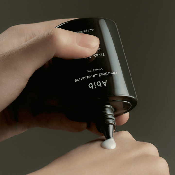 Ruka, ktorá vytláča Abib Heartleaf Sun Essence Calming Drop na vrch druhej ruky, s tmavým pozadím a jasným zobrazením konzistencie produktu.