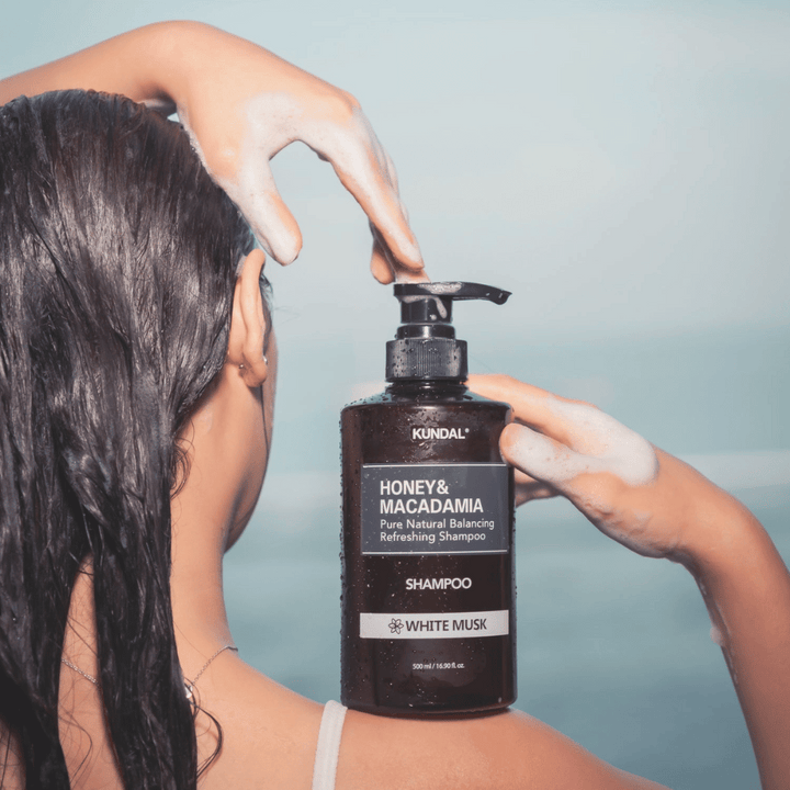 Žena s mokrými vlasmi nanáša penu a drží KUNDAL Honey & Macadamia Nature Shampoo White Musk s pozadím mora.