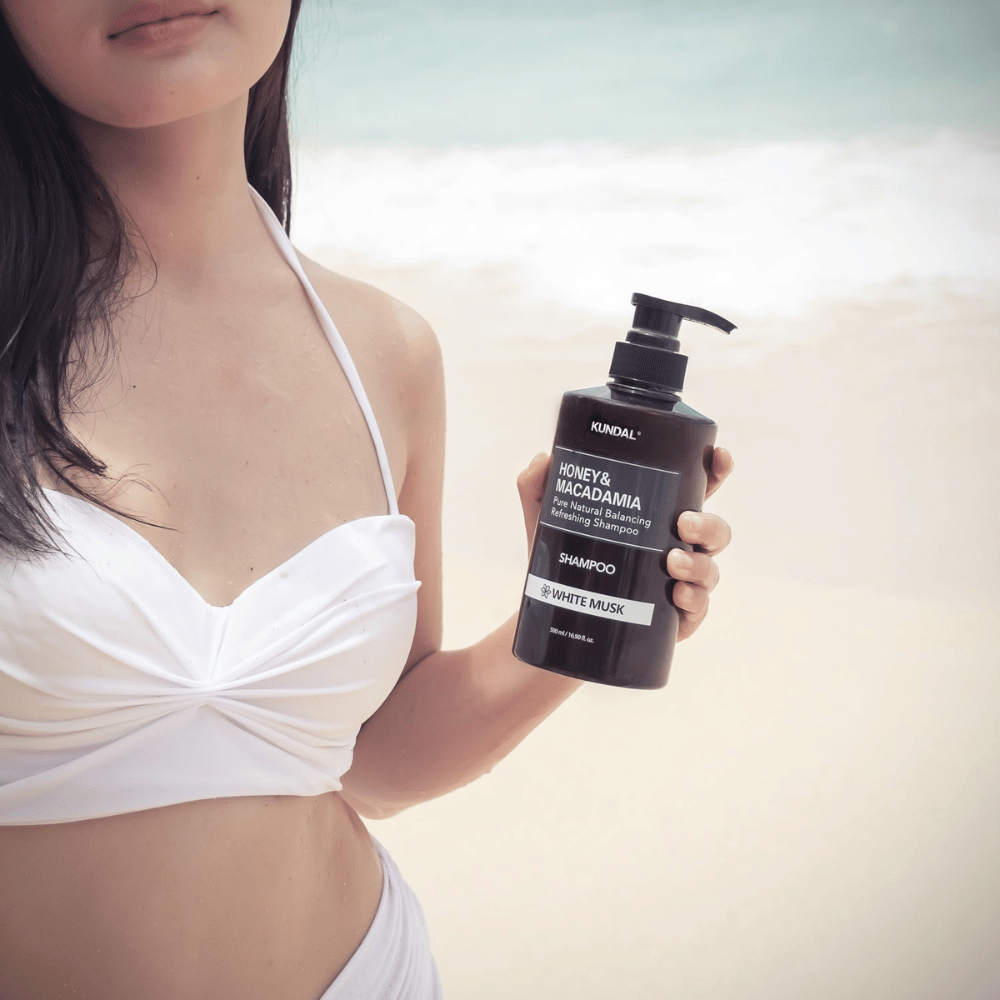 Žena v bielych bikinách drží KUNDAL Honey & Macadamia Nature Shampoo White Musk na pláži s morom v pozadí.