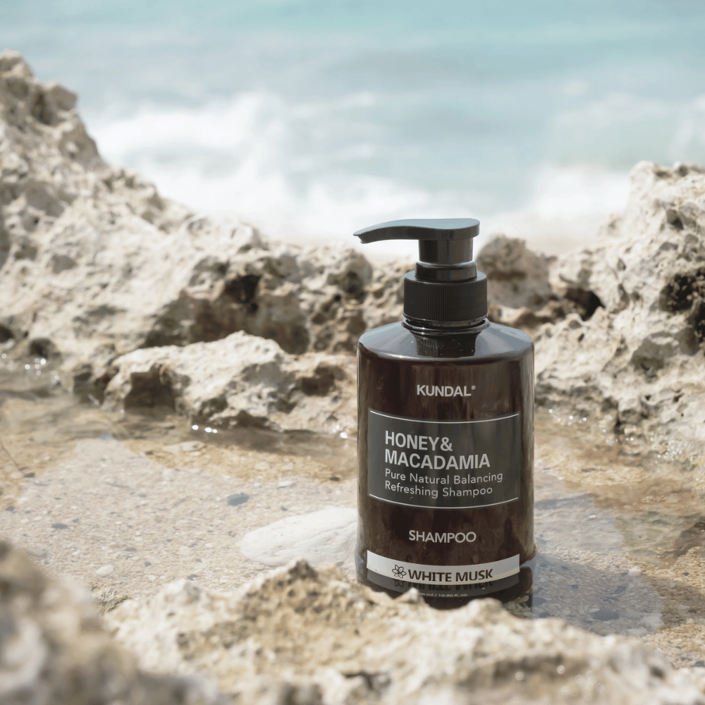 KUNDAL Honey & Macadamia Nature Shampoo White Musk umiestnený na skalách pri mori, prírodný vlasový produkt so sviežou vôňou.