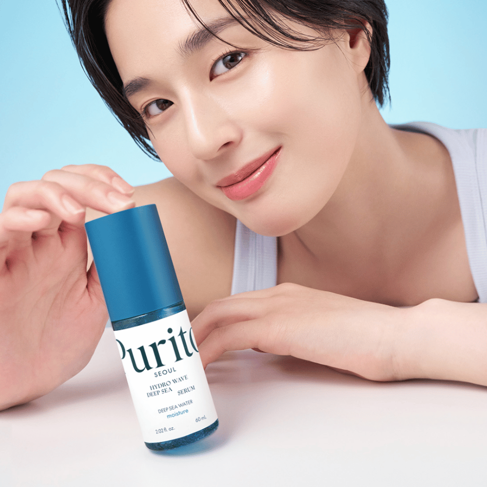 Žena s krátkymi vlasmi drží modré sérum od Purito, Hydro Wave Deep Sea Serum. Jej pleť vyzerá žiarivo a hydratovane, čo zdôrazňuje produkt v centre pozornosti. Pozadie je svetlomodré, čo dodáva svieži a čistý pocit. Produkt je zameraný na starostlivosť o pleť.