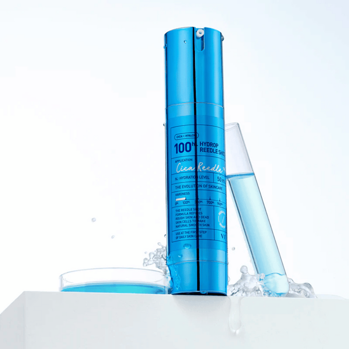 Modrá fľaša VT Cica Hyalon Hydrop Reedle Shot 100hL stojaca na bielom povrchu s vodnými kvapkami, vedľa skúmavky naplnenej modrou tekutinou a okrúhlym viečkom s rovnakou tekutinou.