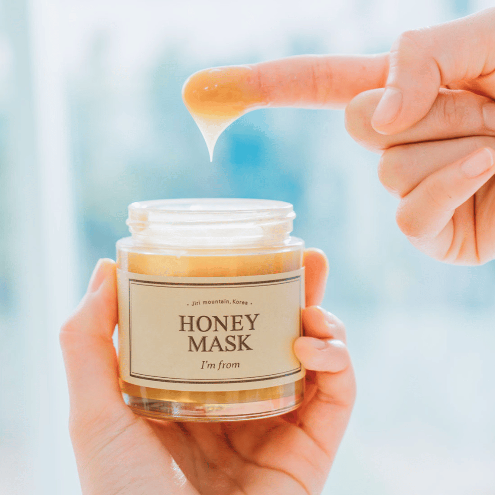 Ruka drží otvorenú plechovku "I'm from Honey Mask" a prst sa ponára do zlatej masky proti svetlému pozadiu.