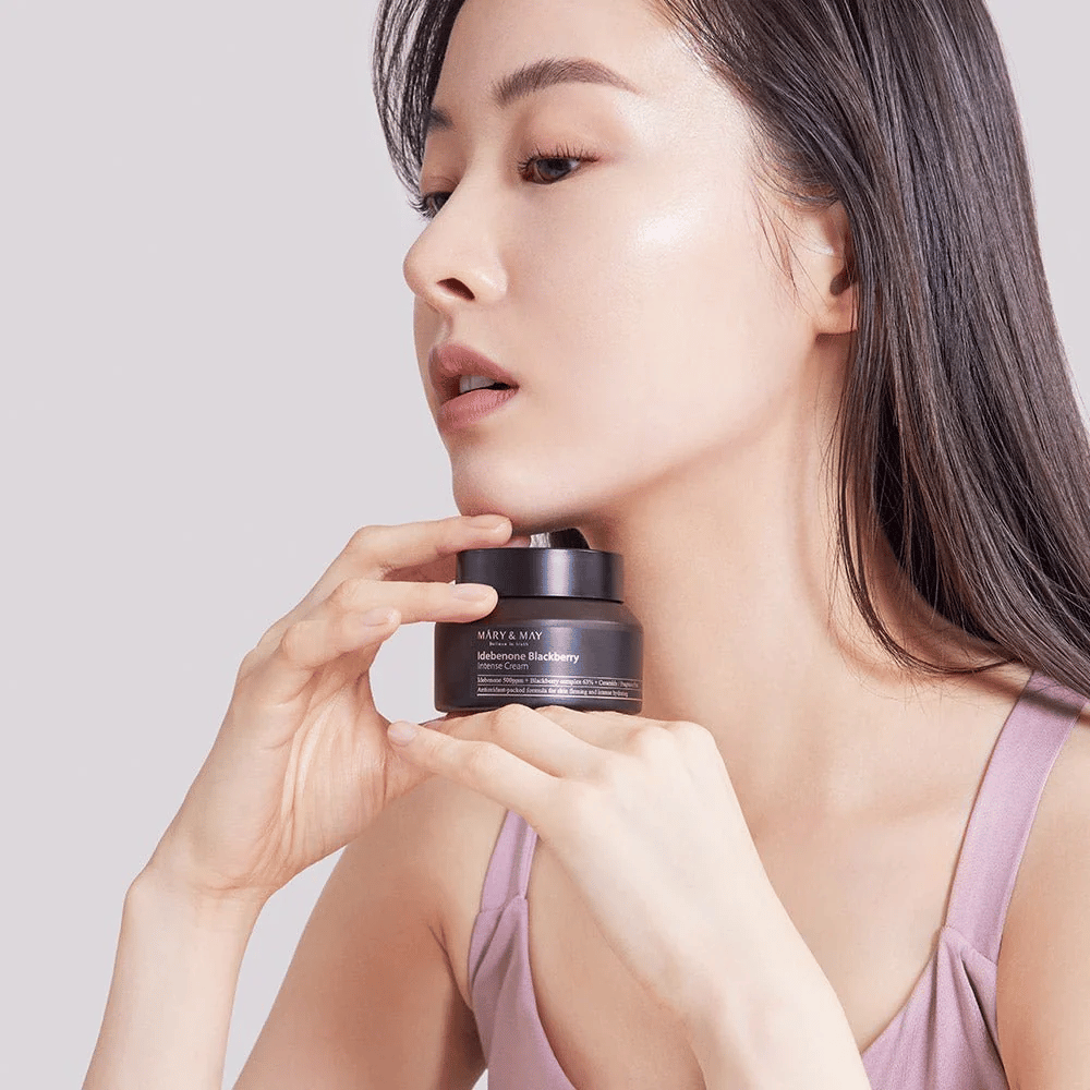 Žena drží pohár s MARY & MAY Idebenone Blackberry Intense Cream. Má žiarivú pleť a jemne sa usmieva, oblečená v tielku. Produkt je intenzívny krém s idebenónom a komplexom černíc, určený na pevnosť a hydratáciu pokožky.