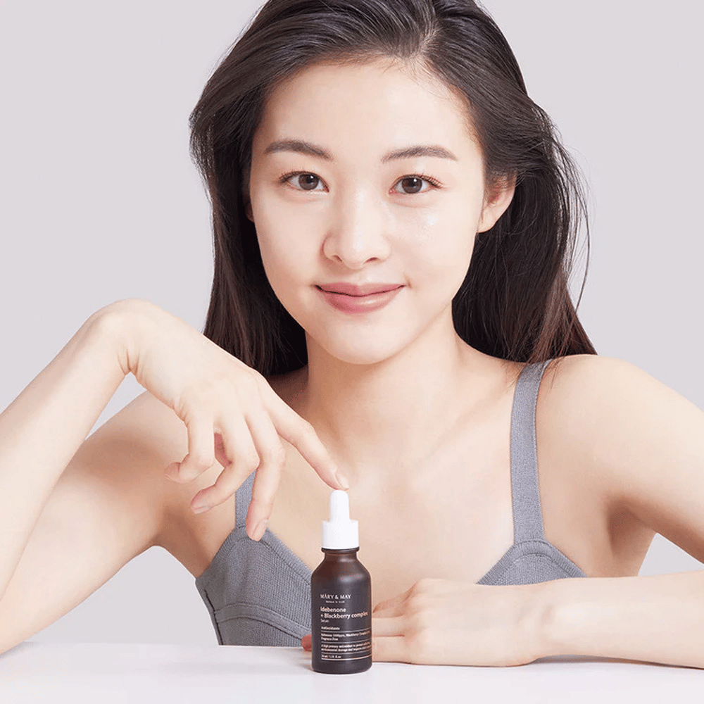 Žena s dlhými, tmavými vlasmi drží sérum MARY & MAY Idebenone + Blackberry Complex, tmavú sklenenú fľašu s pipetou. Má veselý úsmev a ukazuje na fľašu druhou rukou. Žena je oblečená v šedom, bezrukávovom tope a zdá sa, že sa nachádza v svetlom a jednoduchom prostredí, čo zdôrazňuje zameranie produktu na starostlivosť o pleť a krásu.