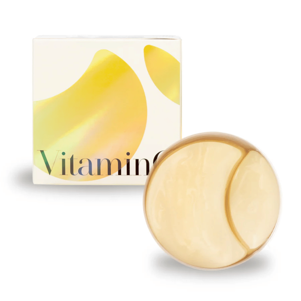 K-Secret Extra Illuminating Eye Gel Patches s vitamínom C v zlatej trblietavej hydrogélovej forme, pohár a balenie v pozadí.