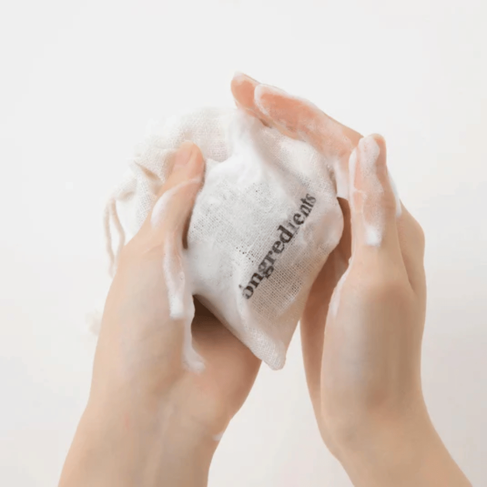 Ruky, ktoré napenívajú Óngredients Jeju Green Tea Cleansing Ball v textilnom vrecku – vegánske čistenie tváre so zeleným čajom.