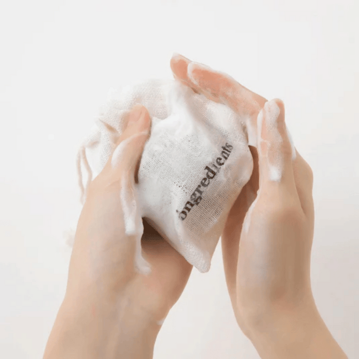 Ruky, ktoré napenívajú Óngredients Jeju Green Tea Cleansing Ball v textilnom vrecku – vegánske čistenie tváre so zeleným čajom.