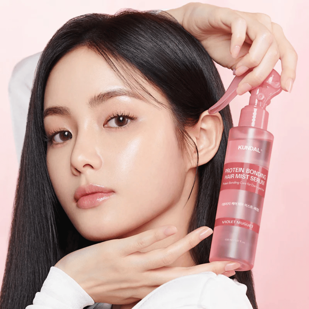 Žena s dlhými čiernymi vlasmi drží KUNDAL Protein Bonding Hair Mist Serum Violet Muguet pred tvárou, na svetloružovom pozadí.