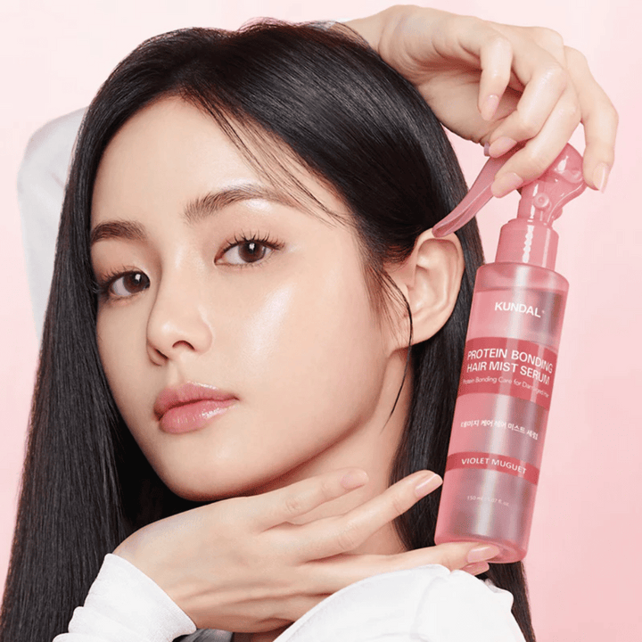 Žena s dlhými čiernymi vlasmi drží KUNDAL Protein Bonding Hair Mist Serum Violet Muguet pred tvárou, na svetloružovom pozadí.
