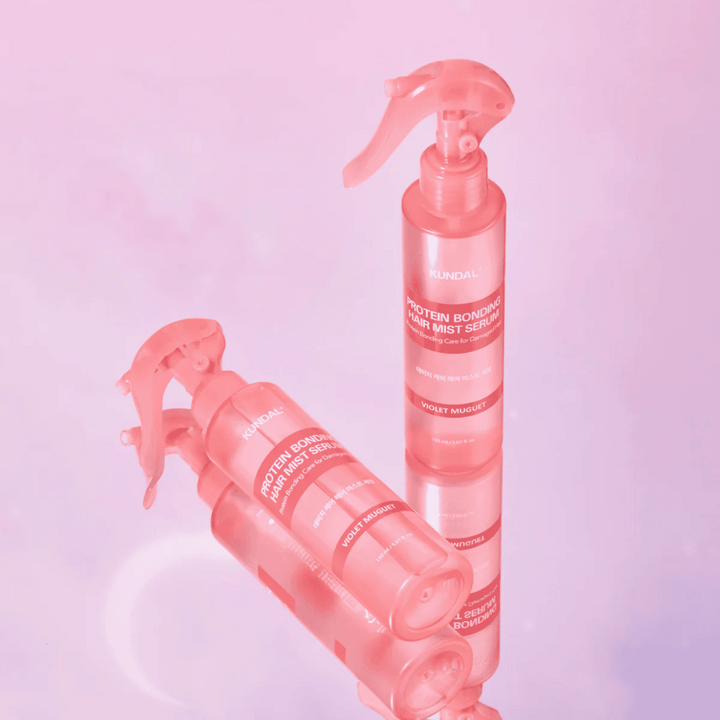 KUNDAL Protein Bonding Hair Mist Serum Violet Muguet v ružových sprejových fľašiach, stojace a ležiace na svetloružovom pozadí s odrazom.