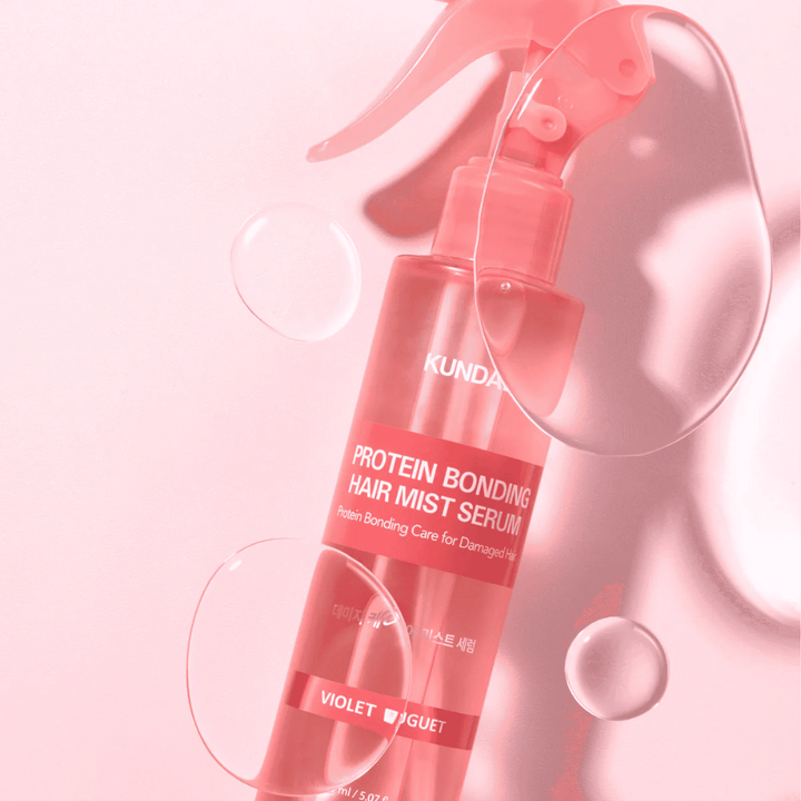 KUNDAL Protein Bonding Hair Mist Serum Violet Bouquet v ružovej sprejovej fľaši, fotografované na ružovom pozadí s kvapkami vody.
