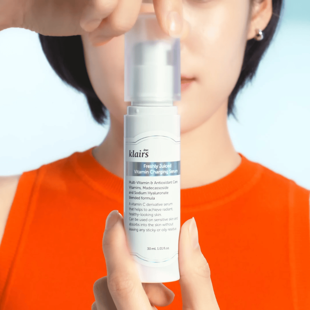 Žena v oranžovom tričku drží Klairs Freshly Juiced Vitamin Charging Serum pred sebou na modrom pozadí.
