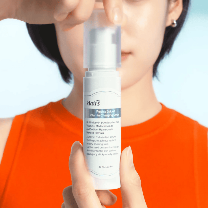 Žena v oranžovom tričku drží Klairs Freshly Juiced Vitamin Charging Serum pred sebou na modrom pozadí.