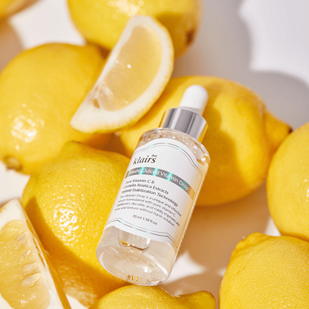Fľaša séra Klairs Freshly Juiced Vitamin Drop obklopená čerstvými polovicami citrónov a plátkami citrónov na svetlom povrchu, zdôrazňuje ingrediencie.