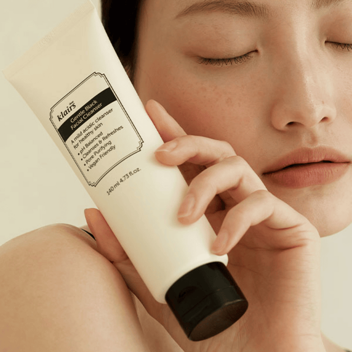 Osoba drží tubu Klairs Gentle Black Facial Cleanser pri svojej tvári, zatvorené oči a prirodzený tón pleti sú viditeľné v pozadí.