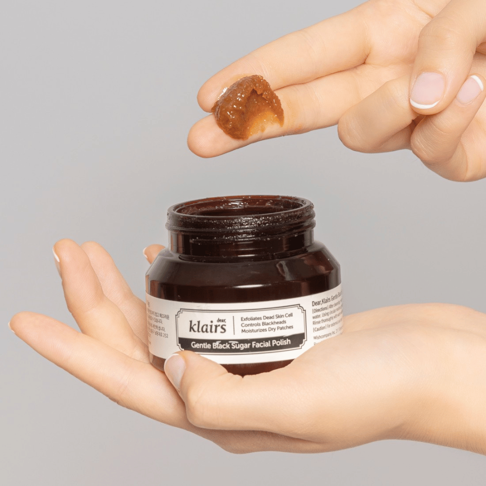 Ruky držia otvorený pohár Klairs Gentle Black Sugar Facial Polish a ukazujú produkt na prste, na šedom pozadí.