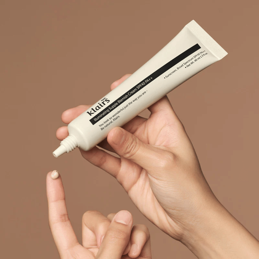 Ruka drží tubu Klairs Illuminating Supple Blemish Cream SPF40 PA++ a vytláča malé množstvo krému na prst, na hnedé pozadie.