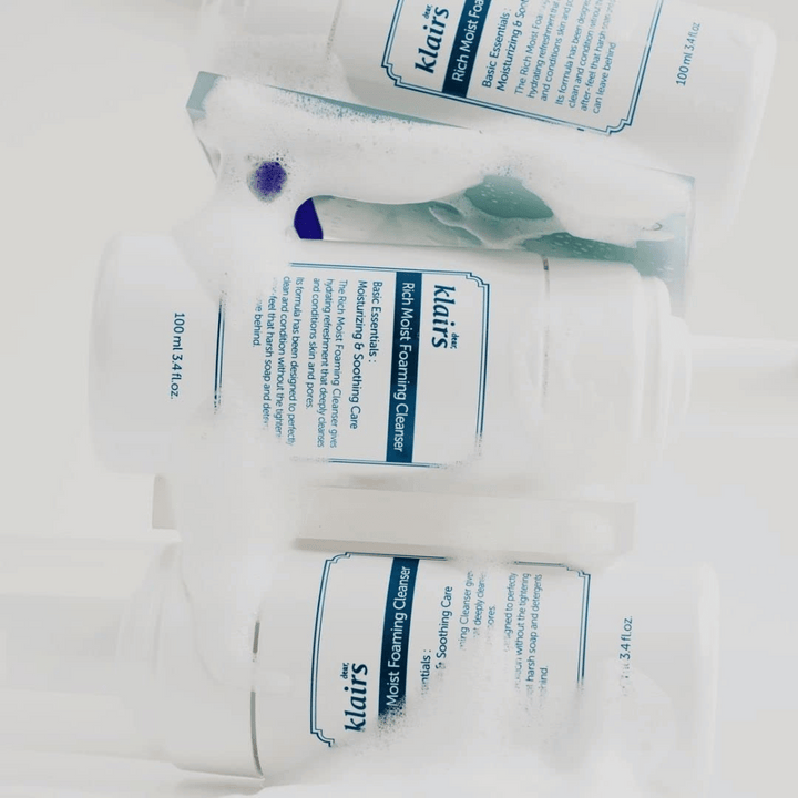 Tri fľaše Klairs Rich Moist Foaming Cleanser pokryté penou a kvapkami vody, umiestnené na bielom povrchu.