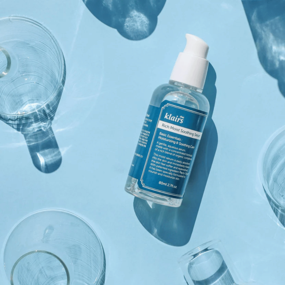 Fľaša Klairs Rich Moist Soothing Serum medzi pohármi vody s odleskami na modrom pozadí.