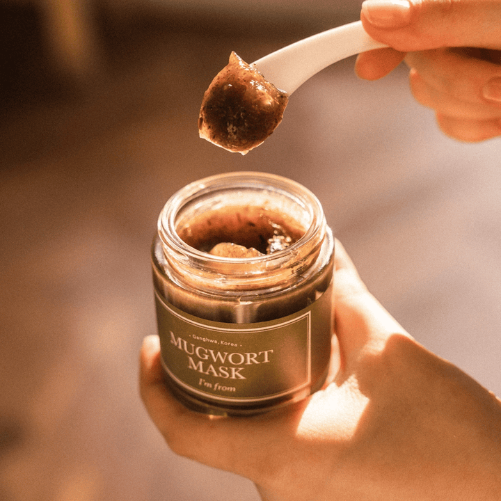 Detailný záber ruky, ktorá drží I'm From Mugwort Mask, zatiaľ čo lyžička naberá bylinkovú gélovú masku z nádoby, v teplom prirodzenom svetle.