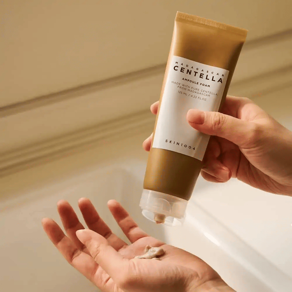 Produkt na obrázku je "Madagascar Centella Ampoule Foam" od SKIN1004. Je to čistiaci prostriedok na tvár vo forme peny, ktorý je vyrobený z Centelly z Madagaskaru. Obsahuje 125 ml / 4.22 fl.oz. Ruka vytláča malé množstvo produktu na použitie.