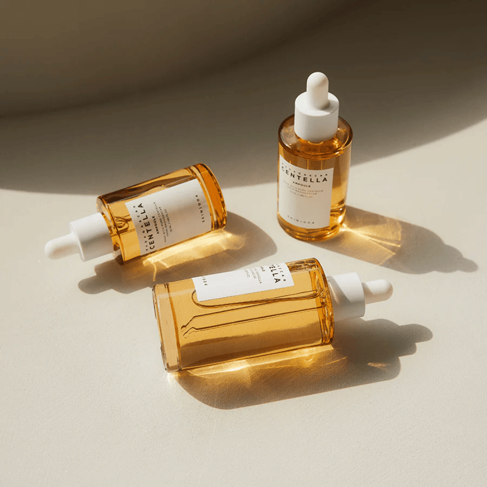 Obrázok ukazuje tri fľaše "Madagascar Centella Asiatica 100 Ampoule". Sú umiestnené na povrchu, kde slnečné svetlo vytvára krásny odraz cez zlatý obsah vo fľašiach. Produkt vyzerá ako súčasť série starostlivosti o pleť, ktorá používa centella asiatica, zložku známu svojimi upokojujúcimi vlastnosťami. Dizajn fliaš je jednoduchý a moderný, čo naznačuje, že ide o prémiovú sériu starostlivosti o pleť.