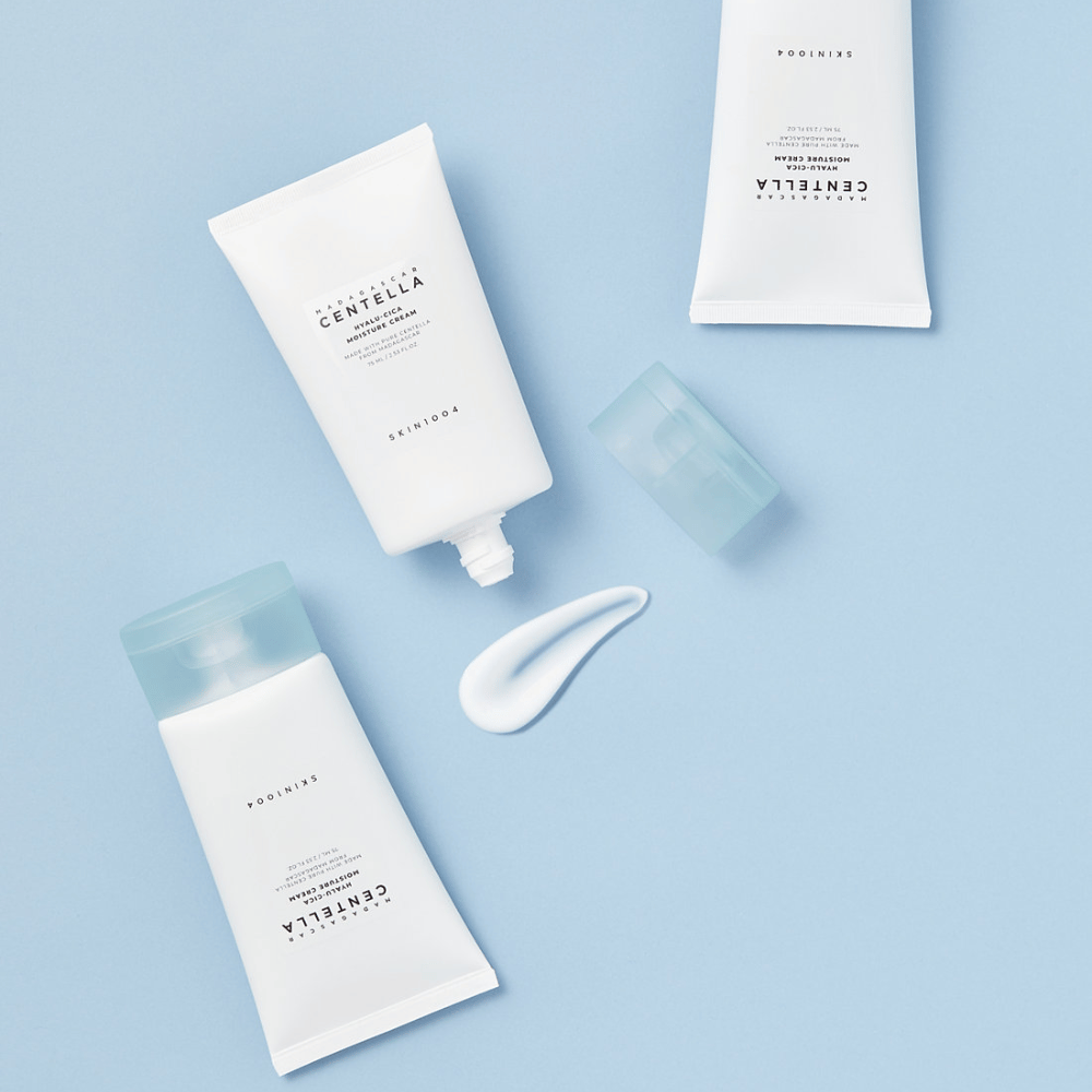 Dve biele tuby s SKIN1004 Centella produktmi starostlivosti o pleť, kvapka produktu a priehľadný modrý uzáver, na svetlomodrom pozadí.