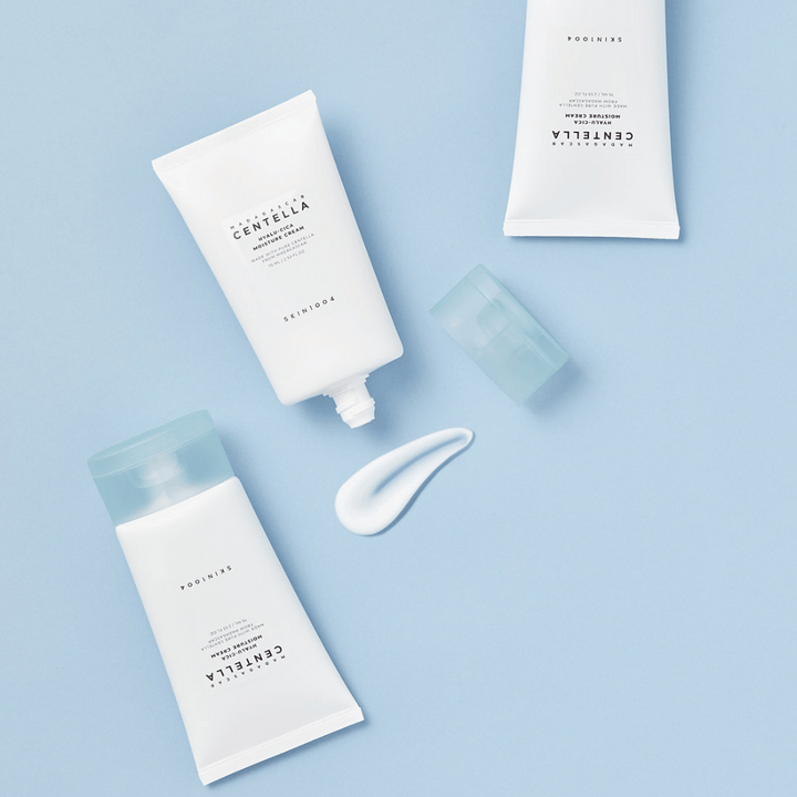 Dve biele tuby s SKIN1004 Centella produktmi starostlivosti o pleť, kvapka produktu a priehľadný modrý uzáver, na svetlomodrom pozadí.