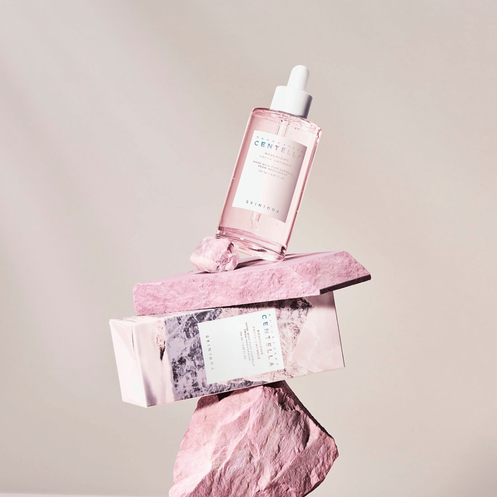 Priehľadná fľaša s ružovou Centella ampulkou od SKIN1004, vyvážená na kamenných blokoch, s kartónovým obalom pod ňou, na neutrálnom pozadí.