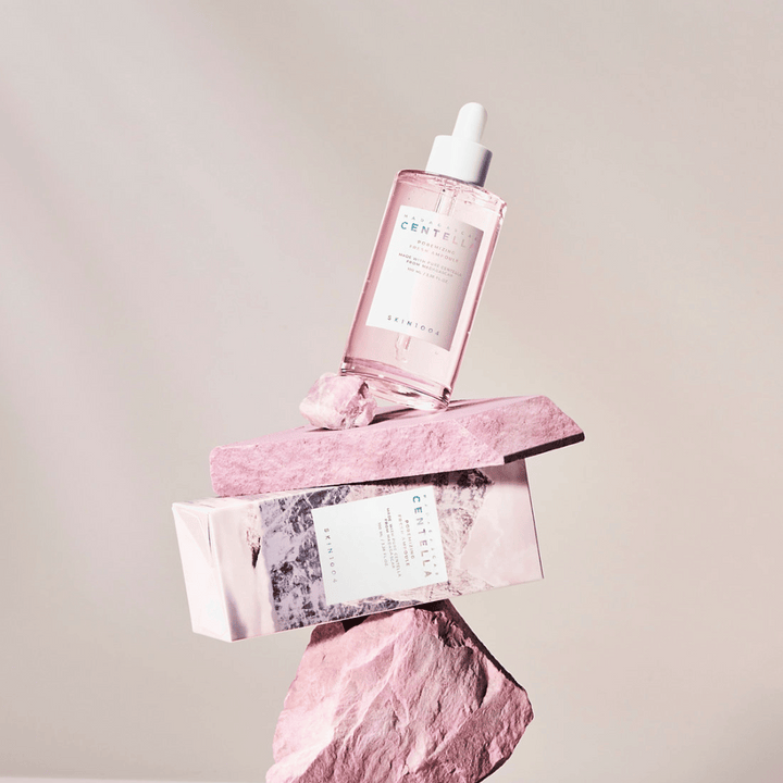 Priehľadná fľaša s ružovou Centella ampulkou od SKIN1004, vyvážená na kamenných blokoch, s kartónovým obalom pod ňou, na neutrálnom pozadí.