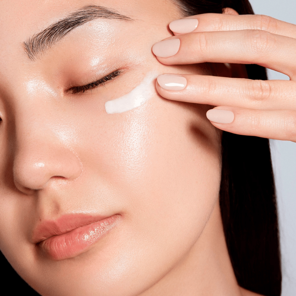 Žena si opatrne nanáša biely očný krém pod oko. Jej pokožka vyzerá žiarivo a hydratovane, čo zdôrazňuje účinok očného krému. Toto vyzdvihuje produkt ako hydratačný a vhodný pre citlivú pokožku okolo očí.