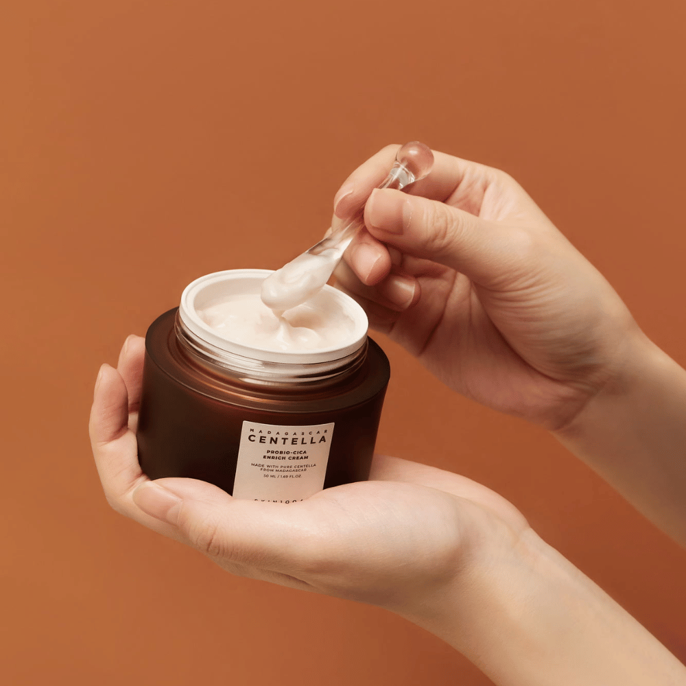 Ruky držiace otvorenú nádobu Madagascar Centella Probio-Cica Enrich Cream a špachtľu s krémom na jednotnom terakotovom pozadí.