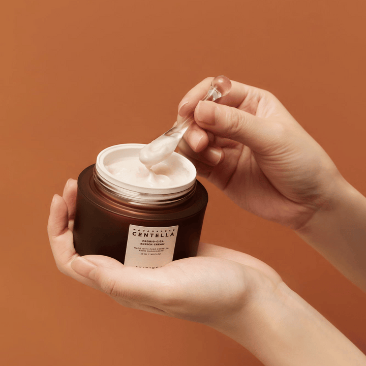 Ruky držiace otvorenú nádobu Madagascar Centella Probio-Cica Enrich Cream a špachtľu s krémom na jednotnom terakotovom pozadí.