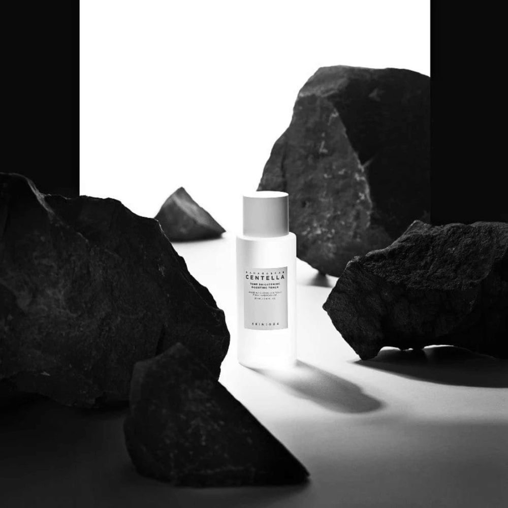 Biela fľaša SKIN1004 Madagascar Centella Tone Brightening Boosting Toner umiestnená dramaticky medzi veľkými čiernymi kameňmi na bielom pozadí.