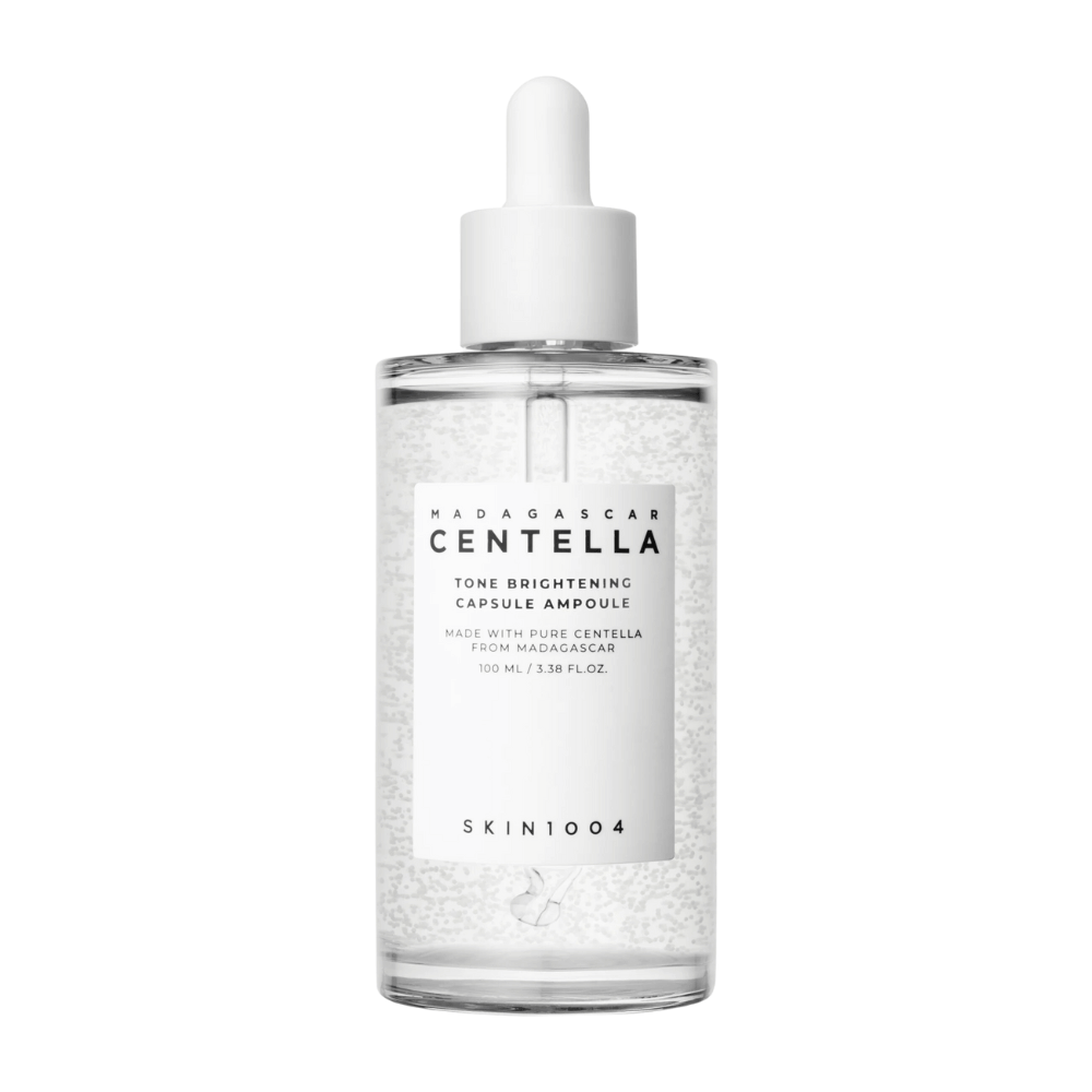 Obrázok zobrazuje fľašu "SKIN1004 Madagascar Centella Tone Brightening Capsule Ampoule". Je to priehľadná fľaša s bielym kvapkadlom. Na etikete je uvedené, že produkt obsahuje čisté centella zložky z Madagaskaru. Obsah fľaše vyzerá, že má malé kapsule alebo častice, čo môže byť súčasťou jej zosvetľujúcich vlastností starostlivosti o pleť. Fľaša obsahuje 100 ml / 3.38 fl.oz. produktu.