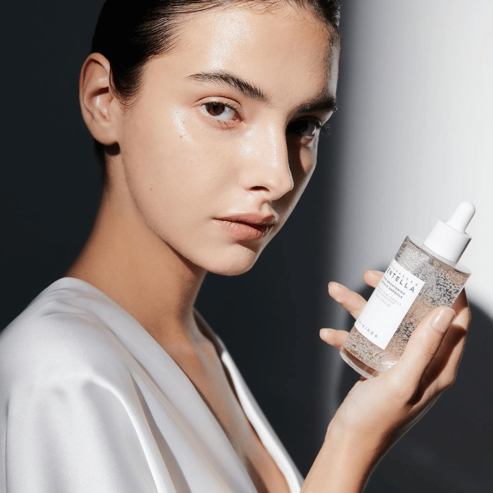 Na obrázku je vidieť osobu, ktorá drží fľašu s kvapkadlom. Fľaša je označená "SKIN1004 Madagascar Centella Tone Brightening Capsule Ampoule". Osoba má žiarivý tón pleti, ktorý odráža svetlo, čo naznačuje možný účinok produktu.