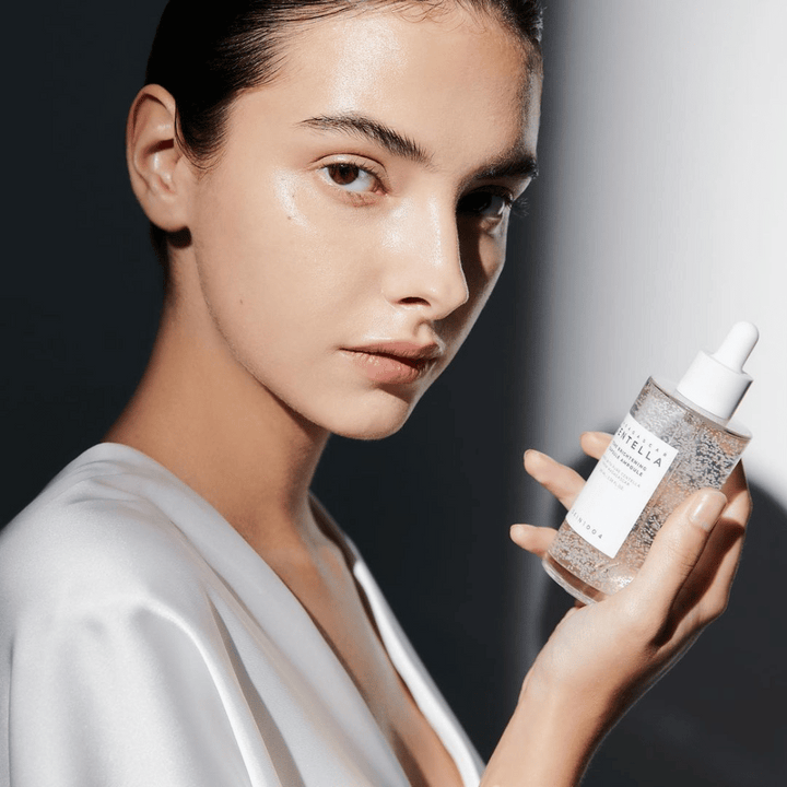 Na obrázku je vidieť osobu, ktorá drží fľašu s kvapkadlom. Fľaša je označená "SKIN1004 Madagascar Centella Tone Brightening Capsule Ampoule". Osoba má žiarivý tón pleti, ktorý odráža svetlo, čo naznačuje možný účinok produktu.