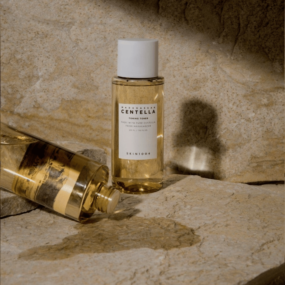 Obrázok zobrazuje fľašu "Madagascar Centella Toning Toner" od SKIN1004. Je to priehľadná fľaša naplnená žltkastou tekutinou, pravdepodobne tonerom na pleťovú starostlivosť. Je uvedené, že je vyrobená s čistou centellou z Madagaskaru a obsah je 210 ml / 7,10 fl.oz. Fľaša a ležiaca fľaša sú umiestnené na kamenistom povrchu, čo vytvára prirodzený a minimalistický pocit.