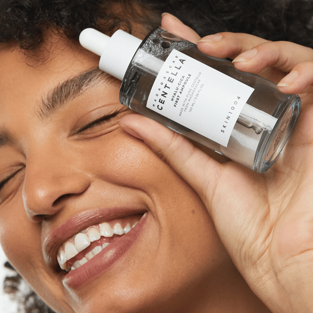 Usmievajúca sa žena drží Skin1004 Madagascar Centella Hyalu-Cica First Ampoule vedľa tváre.
