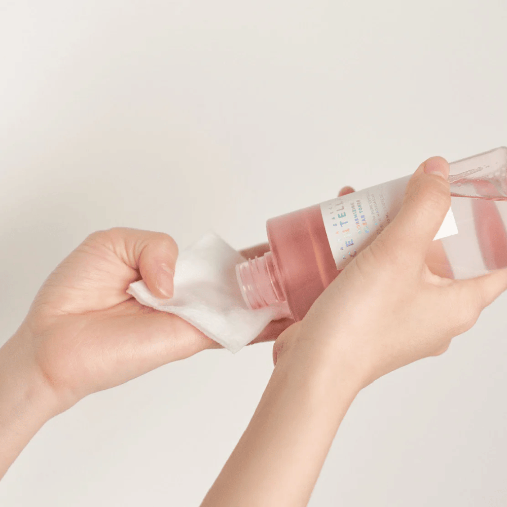 Osoba nalieva Madagascar Centella Poremizing Clear Toner z priehľadnej fľaše na vatový tampón. Produkt, ktorý má ružový odtieň, sa používa na osvieženie a čistenie pleti. Ruky sú viditeľné, čo ukazuje aplikáciu tonera.