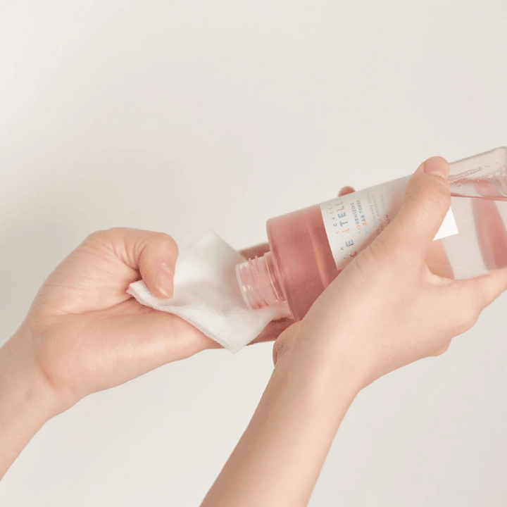 Osoba nalieva Madagascar Centella Poremizing Clear Toner z priehľadnej fľaše na vatový tampón. Produkt, ktorý má ružový odtieň, sa používa na osvieženie a čistenie pleti. Ruky sú viditeľné, čo ukazuje aplikáciu tonera.