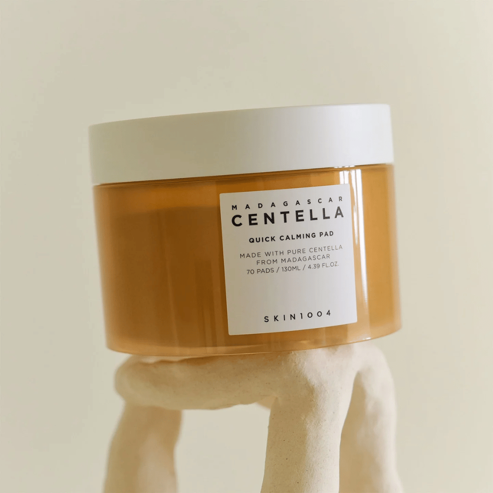 Nádoba s SKIN1004 Madagascar Centella Quick Calming Pad umiestnená na sochársky vyzerajúcom bielom podstavci – jantárová nádoba s bielou etiketou a viečkom.