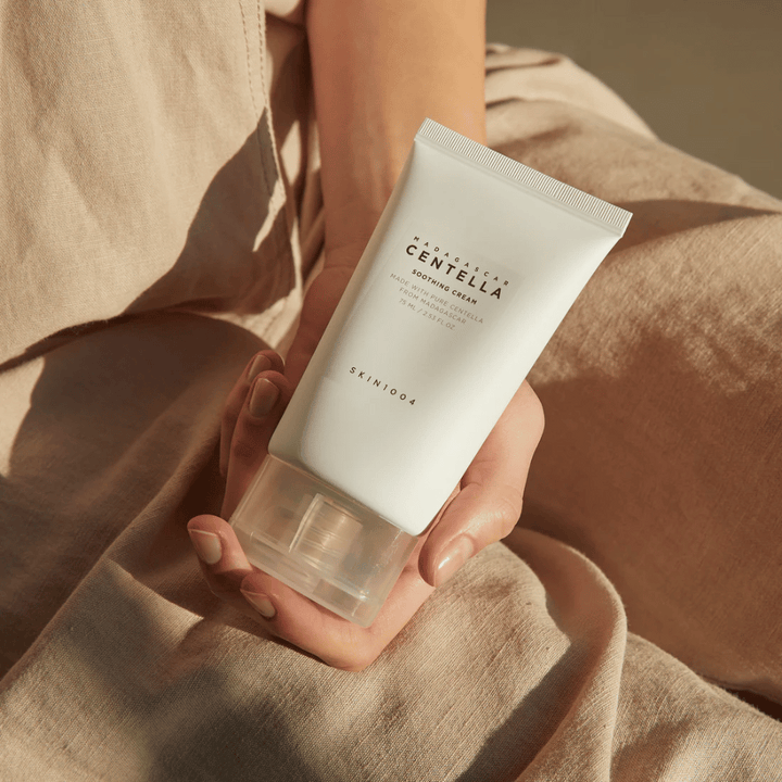 Osoba drží bielu tubu Madagascar Centella Soothing Cream od SKIN1004. Tuba má priehľadný uzáver a text, ktorý opisuje produkt ako upokojujúci a vyrobený z čistej Centelly. Osoba je oblečená v svetlom oblečení.