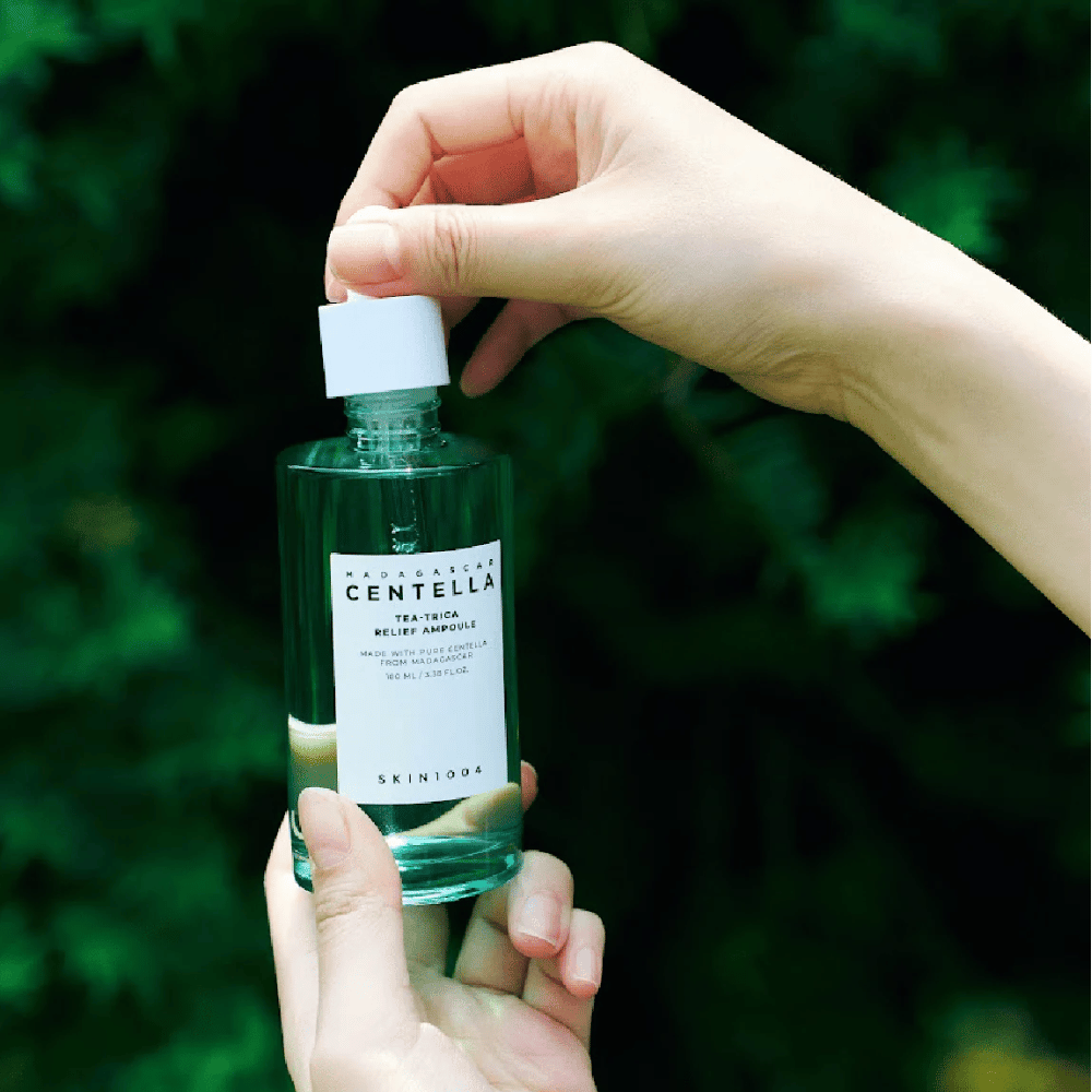 Osoba otvára SKIN1004 Madagascar Centella Tea-Trica Relief Ampoule – zelená sklenená fľaša s bielou etiketou držaná v rukách pred zeleným pozadím.