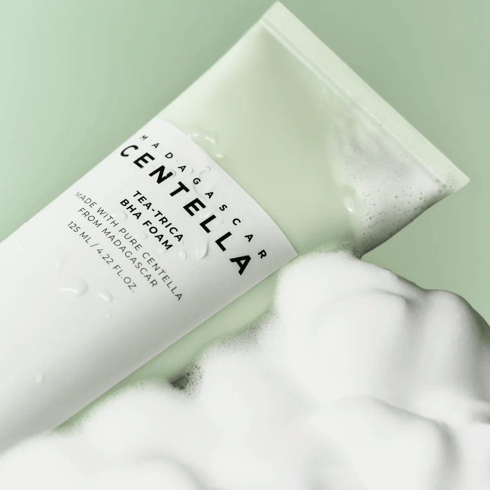 Produkt typu čistiaca pena, "Madagascar Centella Tea-trica BHA Foam," je čiastočne viditeľný s penou okolo. Produkt má zeleno-biely dizajn a texty "Madagascar Centella" a "Tea-trica BHA Foam" sú jasne viditeľné. Objem je 125 ml.