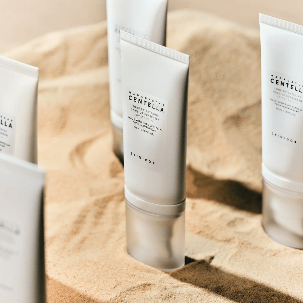 Viaceré tuby s Madagascar Centella Tone Brightening Tone-Up Sunscreen stoja v piesku. Tuba je biela s čiernym textom a obsahuje 50 ml. Produkt má SPF 50+ PA++++ a je vyrobený s čistou Centellou z Madagaskaru.